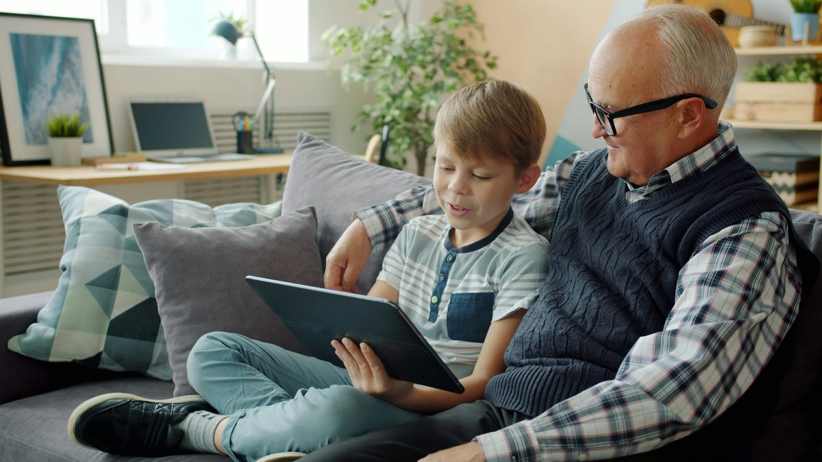 A grandfather and his grandson sitting together on a couch, looking at a tablet and talking